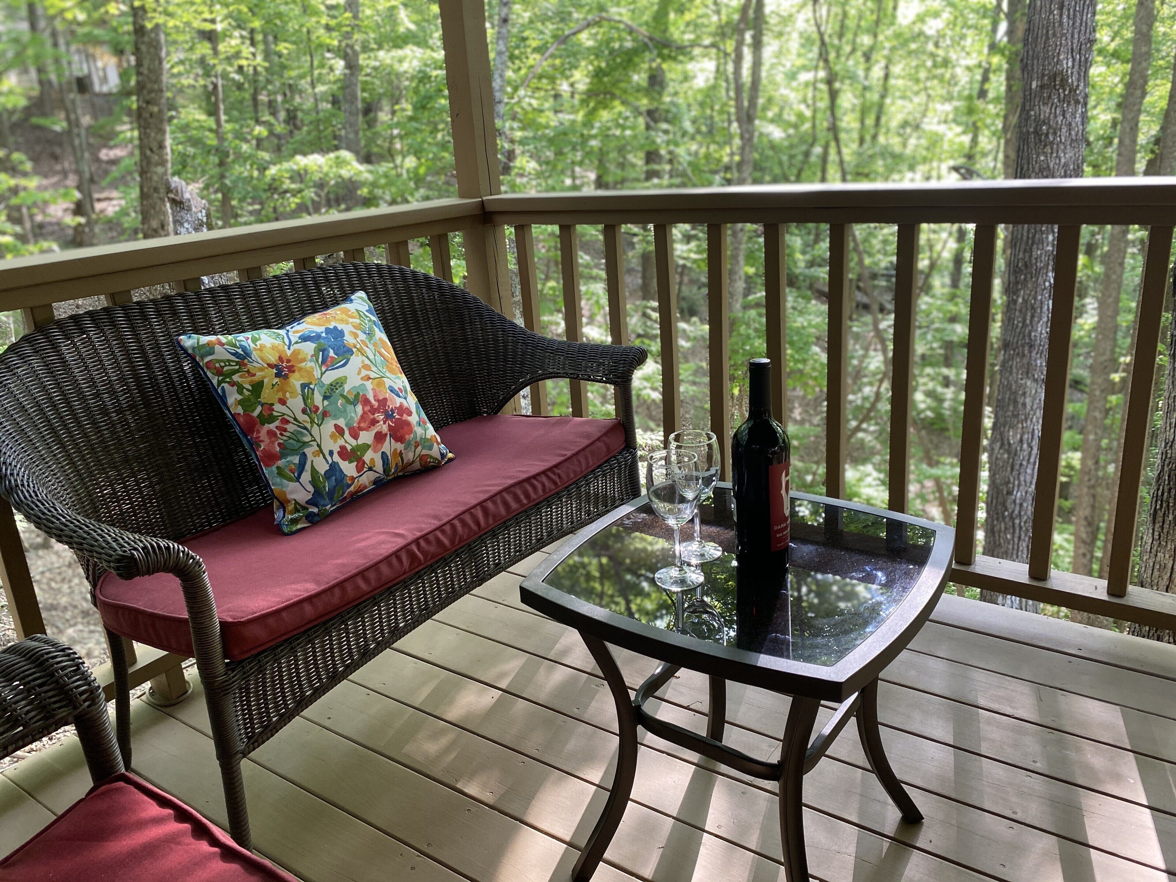 A wicker sofa with a floral cushion and a glass table with a bottle and glasses on a wooden deck with a forest view.