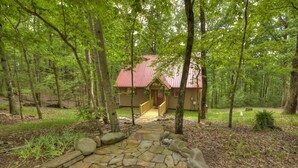 Un chalet rustique avec un toit rouge, entouré d’une forêt dense. Un sentier de pierres mène à l’entrée.