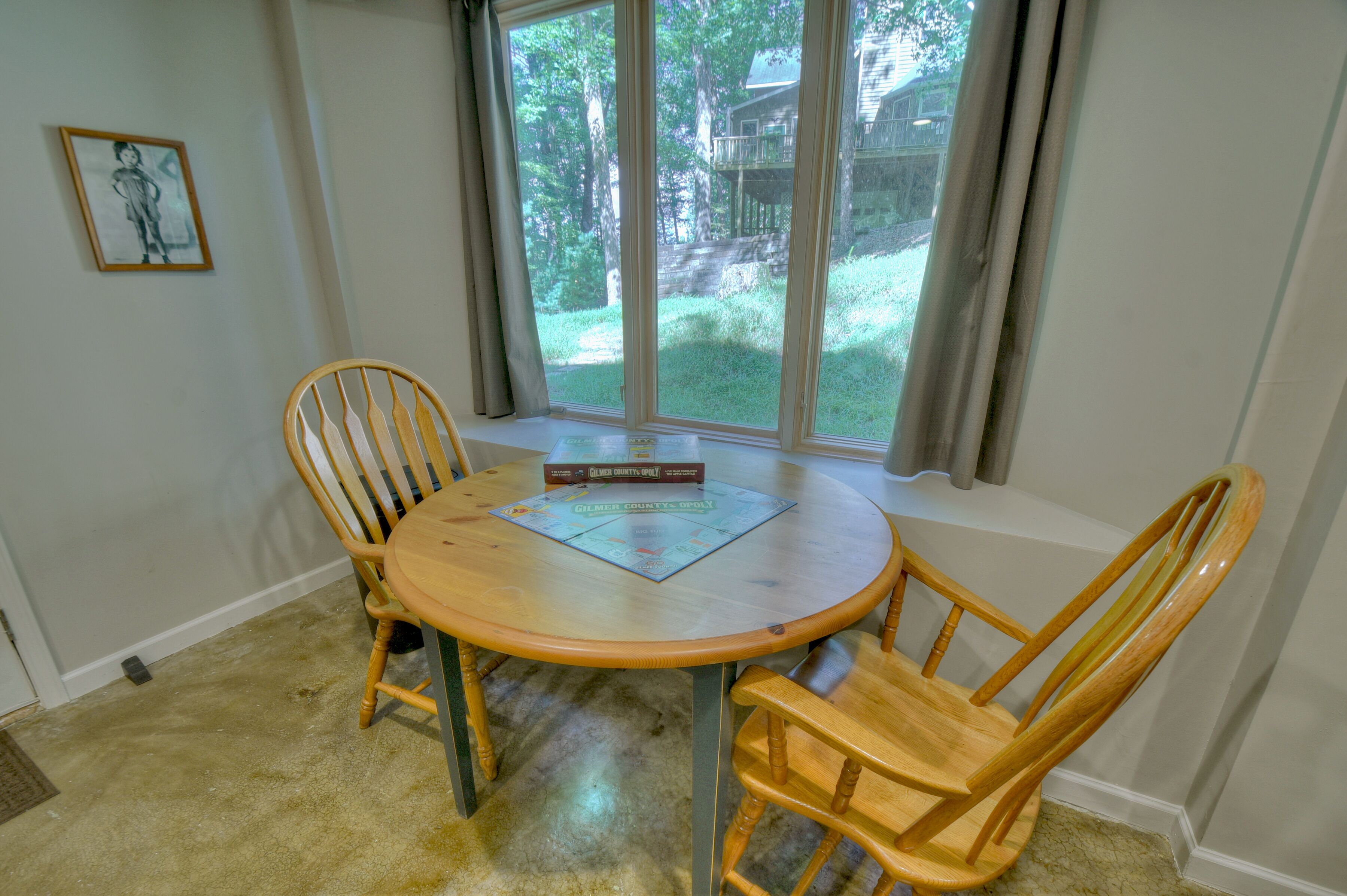 Een eethoek met een ronde houten tafel en vier stoelen, een ingelijst beeld aan de muur en een raam met gordijnen.