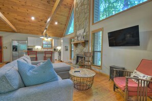 A spacious living area with a wooden ceiling, a stone fireplace, a television, and a kitchen in the background. - Mountain Escape - Hot Tub, Fireplace & Pet Friendly (Ellijay)