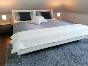 Een net opgemaakt bed met een witte deken en kussens, een grijs tapijt en een bedlampje.