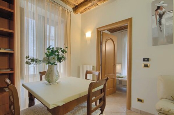 A dining area with a table, chairs, a vase with flowers, a wooden bookshelf, a radiator, and a door leading to another room.