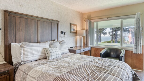 A bedroom with a bed, a nightstand, a chair, a window with a view, and a wall-mounted lamp. - T11 - Trawler @ Trollers Lodge (Depoe Bay)
