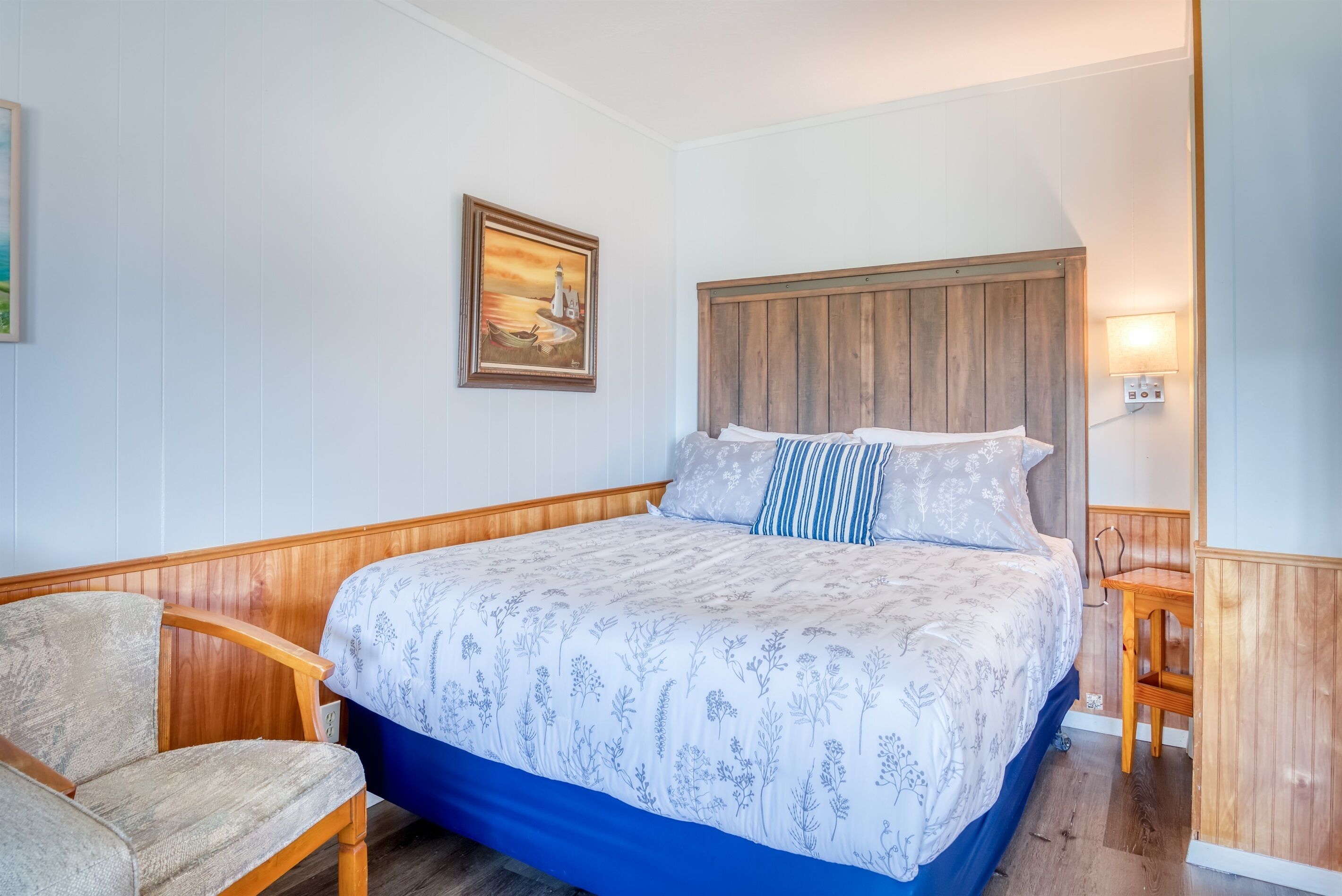 A bedroom with a wooden headboard, a bed with floral bedding, a chair, a painting on the wall, and a nightstand with a lamp.