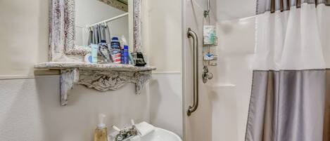 Une salle de bain avec un lavabo blanc, un miroir doté d’un cadre décoratif, une douche équipée d’une barre d’appui et un porte-serviettes.