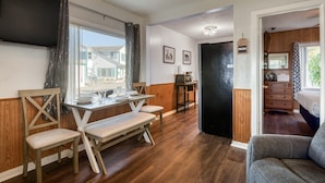 Uma sala de jantar com mesa e cadeiras de madeira, uma cozinha com geladeira, e um quarto com cama e cômoda.