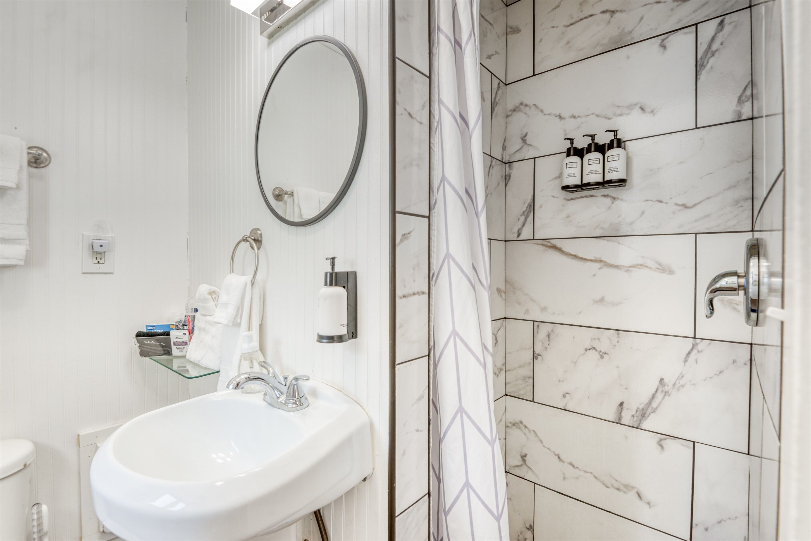 Une salle de bain avec un lavabo blanc, un miroir, une étagère en verre où sont rangés les articles de toilette et une douche aux carreaux de marbre.