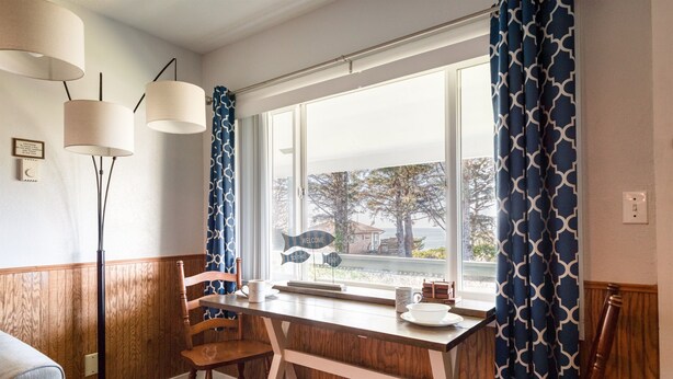 A dining area with a wooden table, chairs, a floor lamp, and a window with a view of trees and a building. - T10 - Skiff @ Trollers Lodge (Depoe Bay)