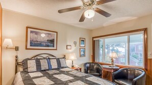 A bedroom with a bed, a ceiling fan, a desk with a chair, a window with a view, and framed pictures on the wall. - T08 - Schooner @ Trollers Lodge (Depoe Bay)