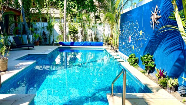 A swimming pool with a blue tiled edge, surrounded by potted plants and a mural on the adjacent wall. - Private home one block to the beach! 2 bedrooms. Premier Zona Dorado location! (Bucerias)