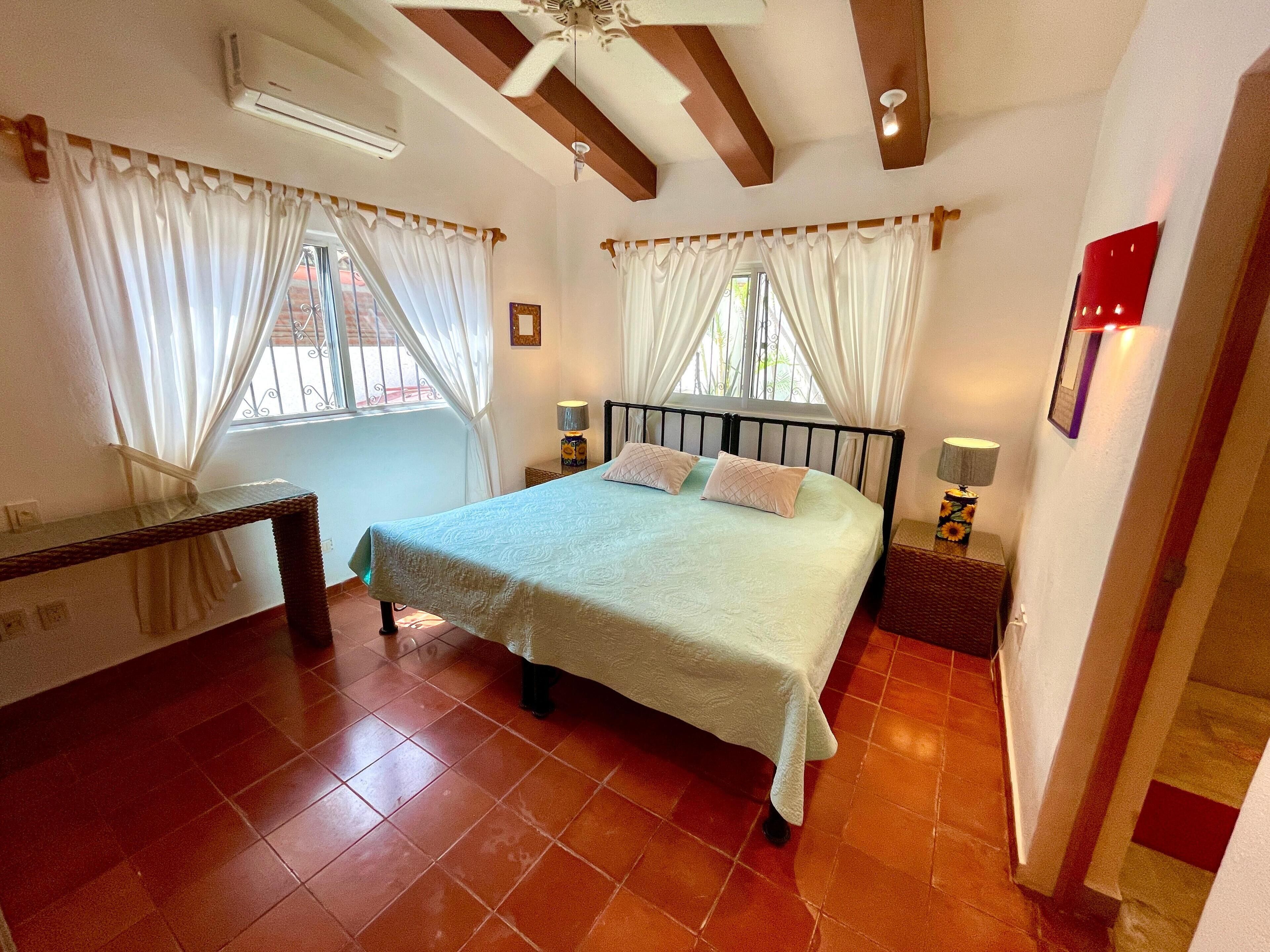 A bedroom with a bed, two bedside tables with lamps, a window with curtains, and a ceiling fan.