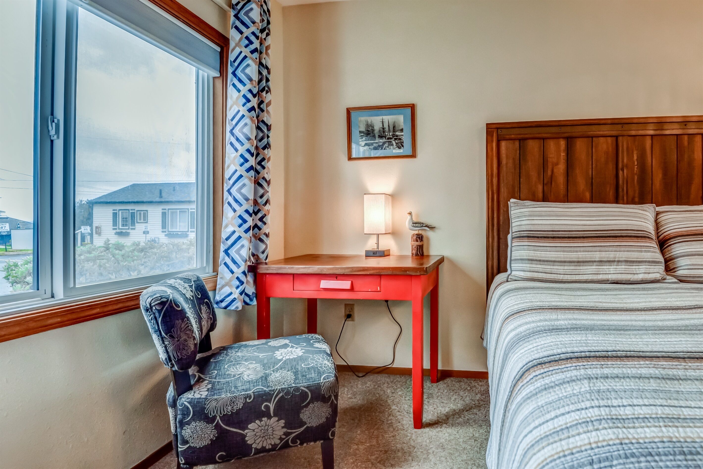 A hotel room with a bed, a desk with a lamp, a chair, a window with a view of houses, and a framed picture on the wall.