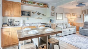 Una cocina compacta con armarios de madera, una mesa de comedor con cuencos blancos y copas de vino, un sofá y un sillón gris en la sala.
