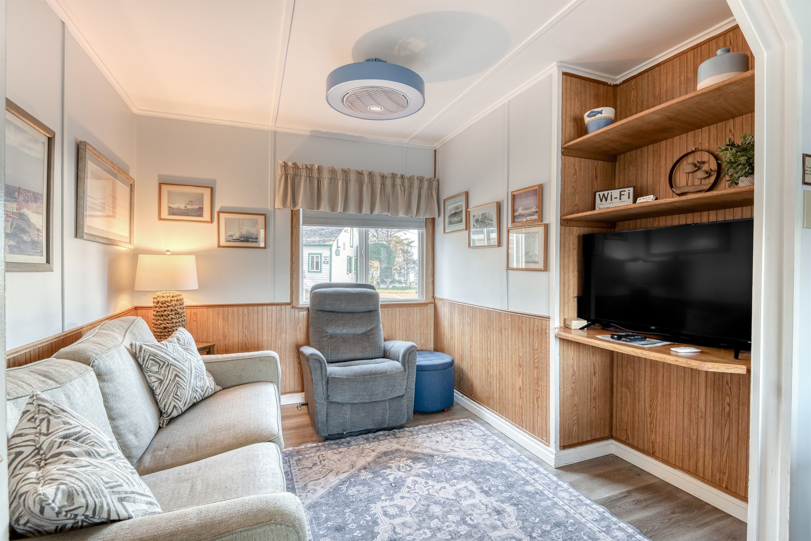 Een woonkamer met een bank, fauteuil, televisie, houten planken en ingelijste foto’s aan de muur.