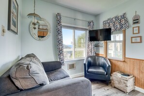 Una habitación con sofá, una silla, televisor, ventana con cortinas y un espejo montado en la pared.
