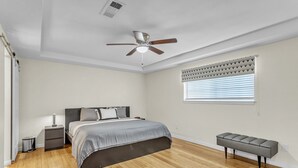 Une chambre à coucher comprenant un lit, une table de chevet, un banc, un ventilateur de plafond, une fenêtre avec stores et un luminaire.