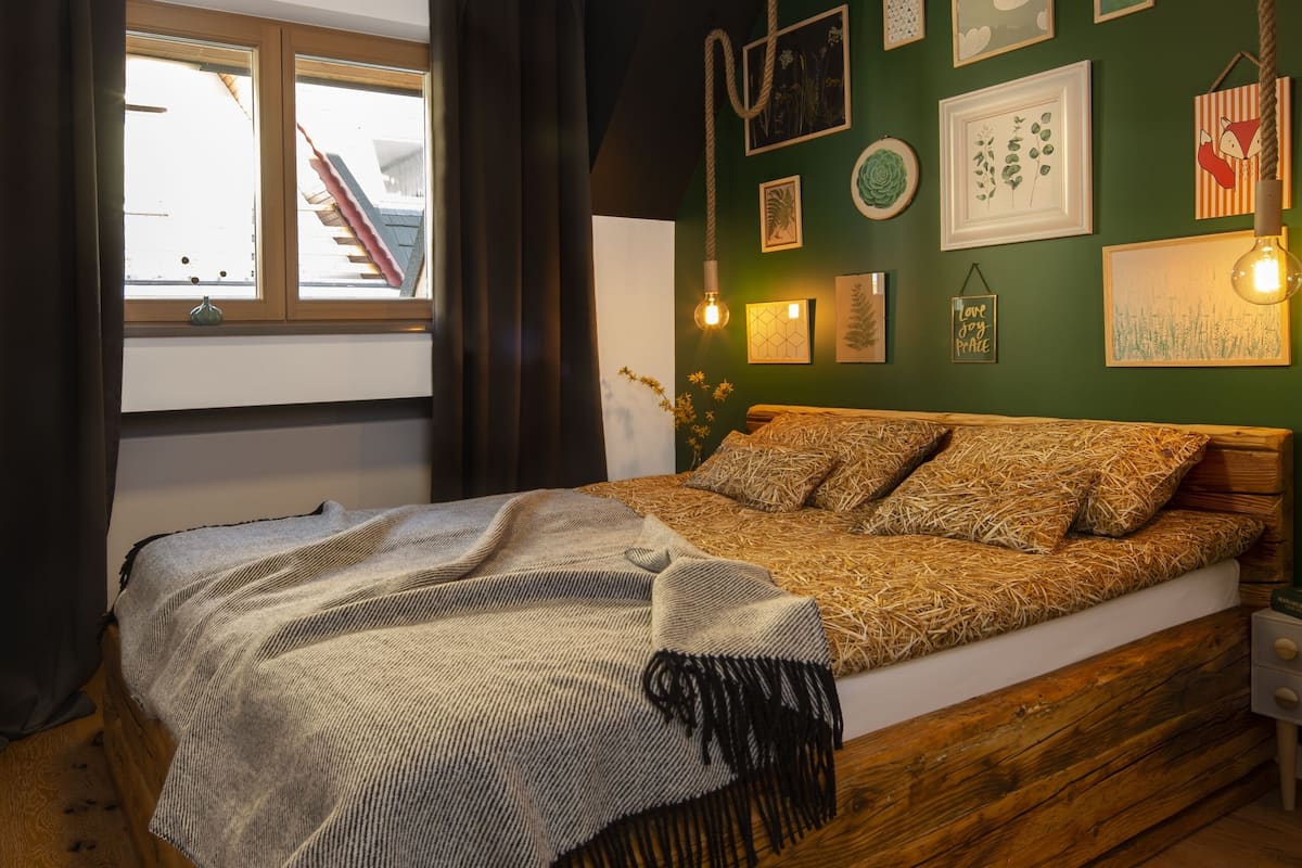 A bedroom with a wooden bed, a green wall with framed pictures, and a window with curtains.