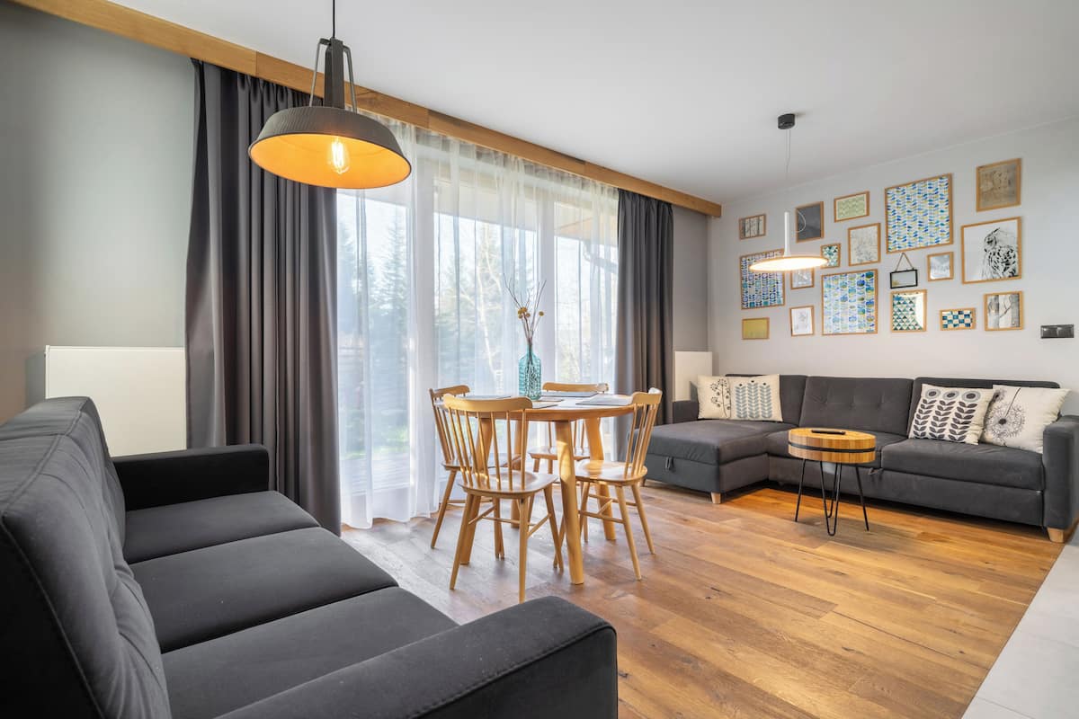 A modern living room with a sofa, dining table, and chairs. There are curtains, a lamp, and framed pictures on the wall.