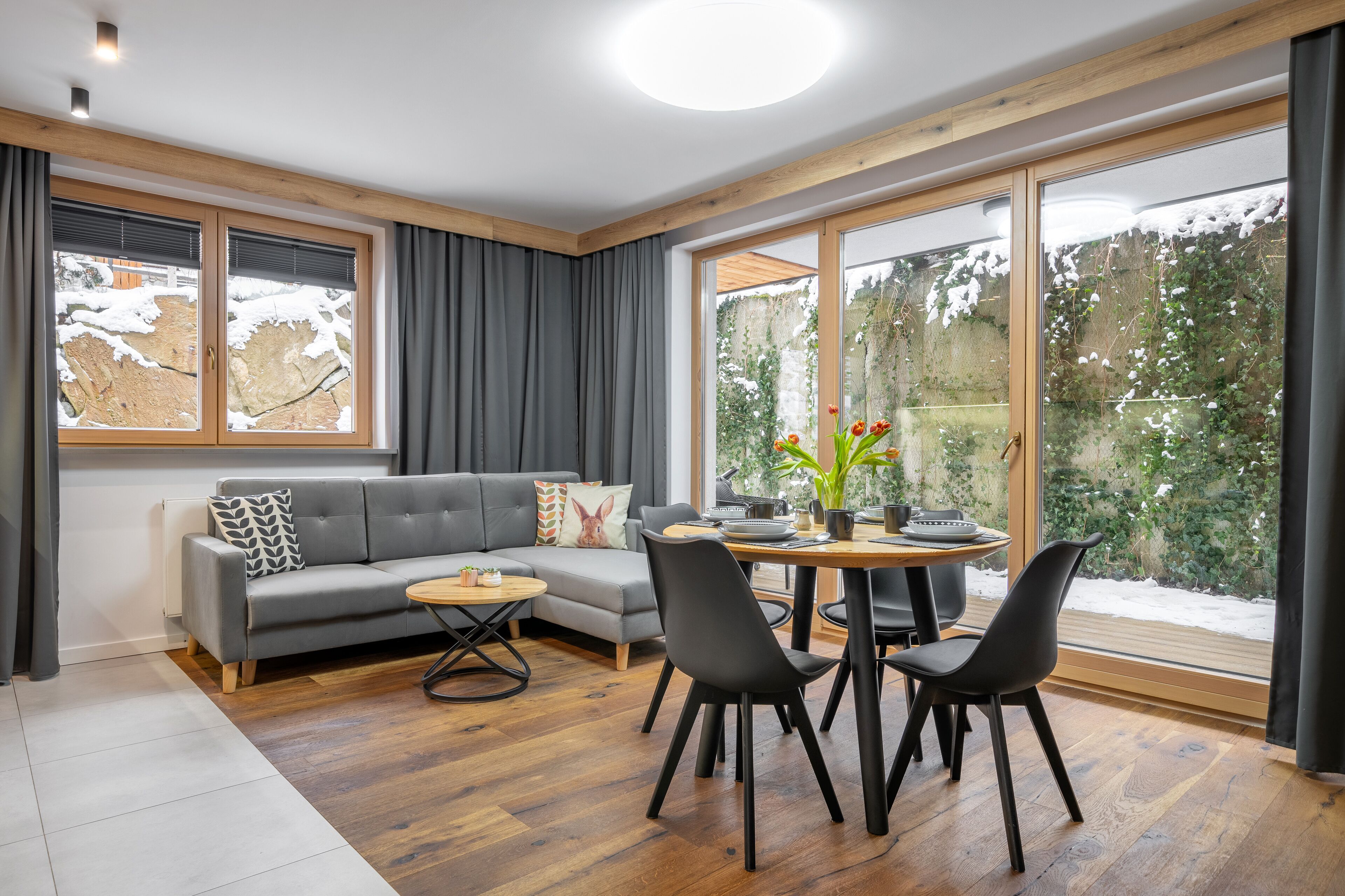A modern living room with a grey sofa, black chairs, a wooden dining table, and large windows with a view of snow-covered trees.