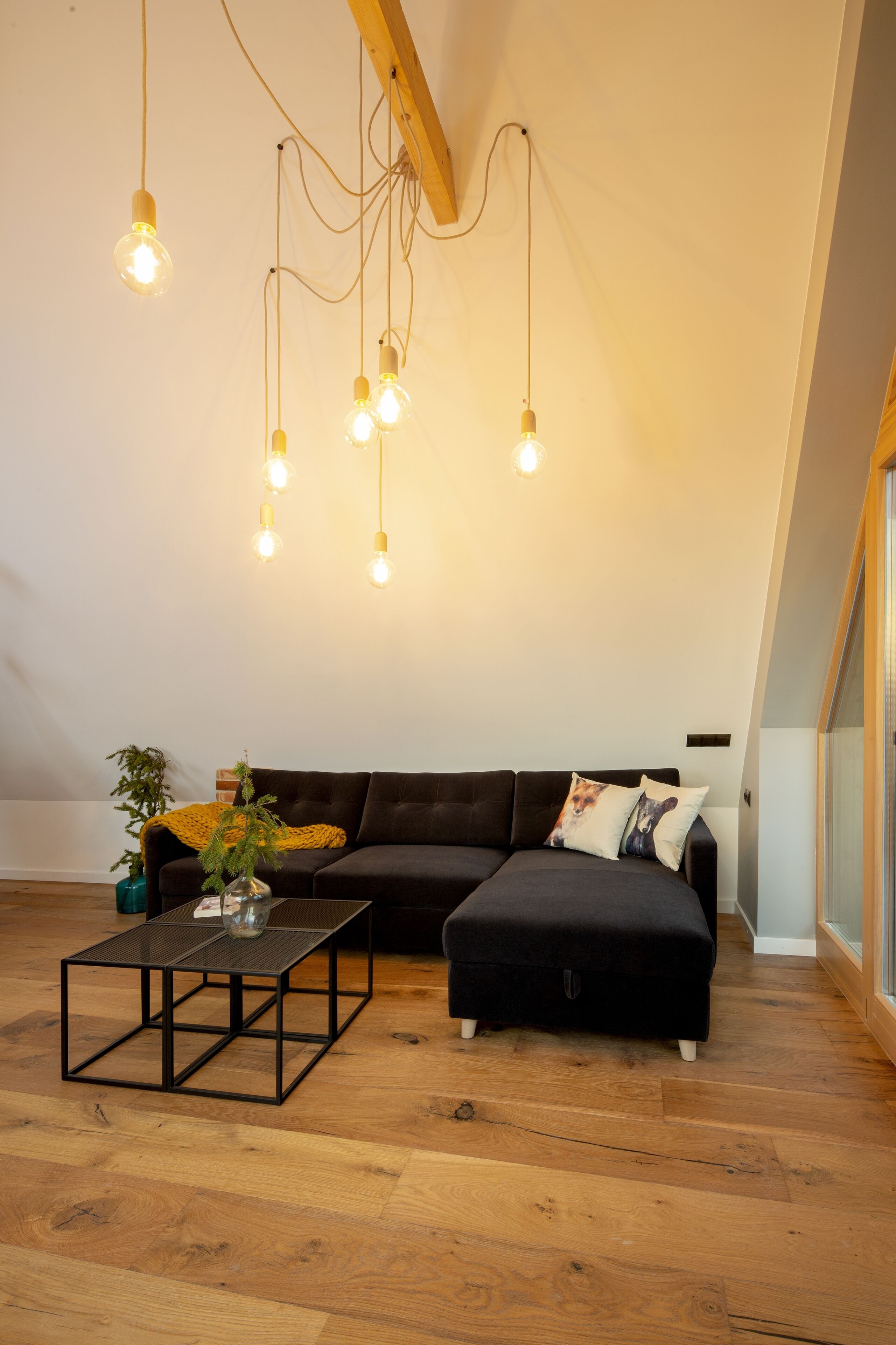 A loft-style room with a dark brown sofa, a black ottoman, a glass coffee table, and a hanging light fixture with multiple bulbs.
