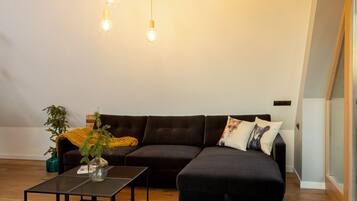 A loft-style room with a dark brown sofa, a black ottoman, a glass coffee table, and a hanging light fixture with multiple bulbs.