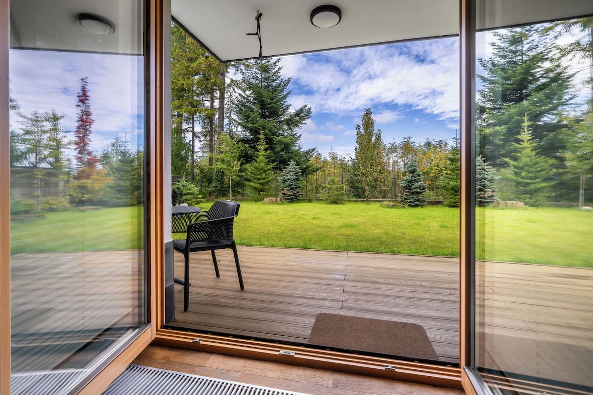 A room with a large glass door leading to an outdoor deck with a wooden floor, a black chair, and a view of a grassy yard with trees.