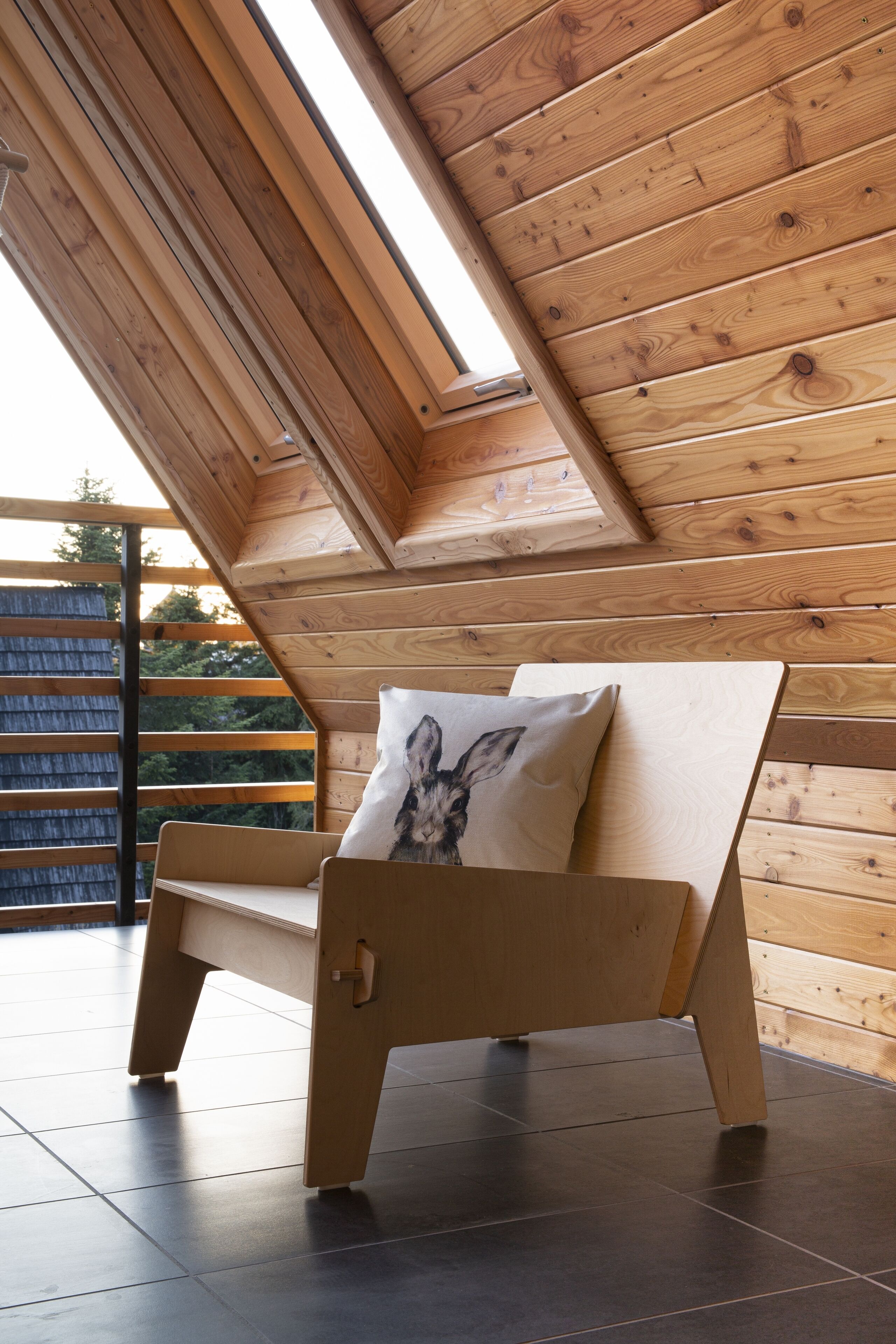 A wooden bench with a cushion in a room with sloped wooden walls and a view of greenery outside.