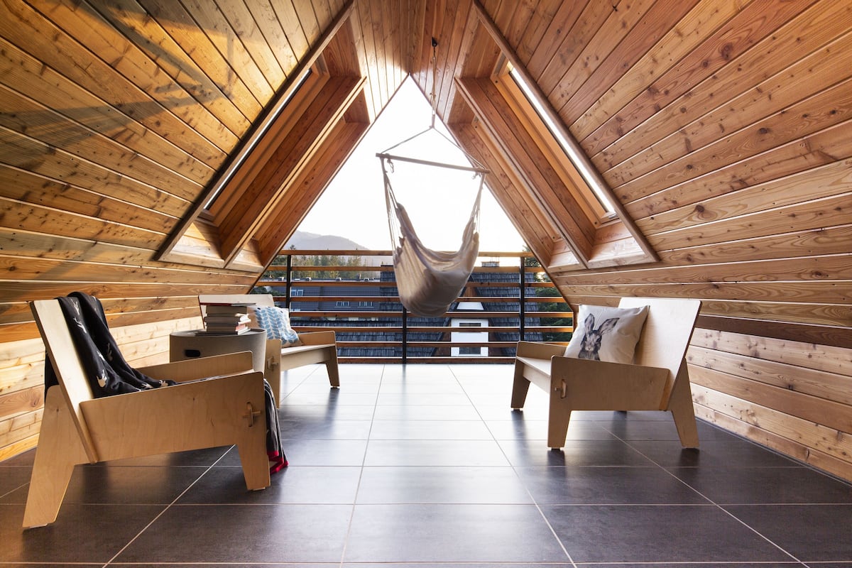 A cozy attic room with wooden walls and a sloped ceiling, featuring a hammock, two wooden benches, and a small table with books and a lamp.