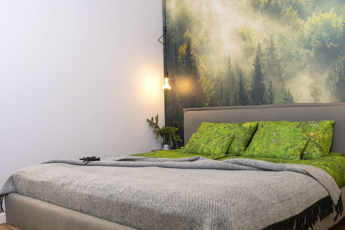 A modern bedroom with a large bed, a wall-mounted lamp, and a forest-themed mural.