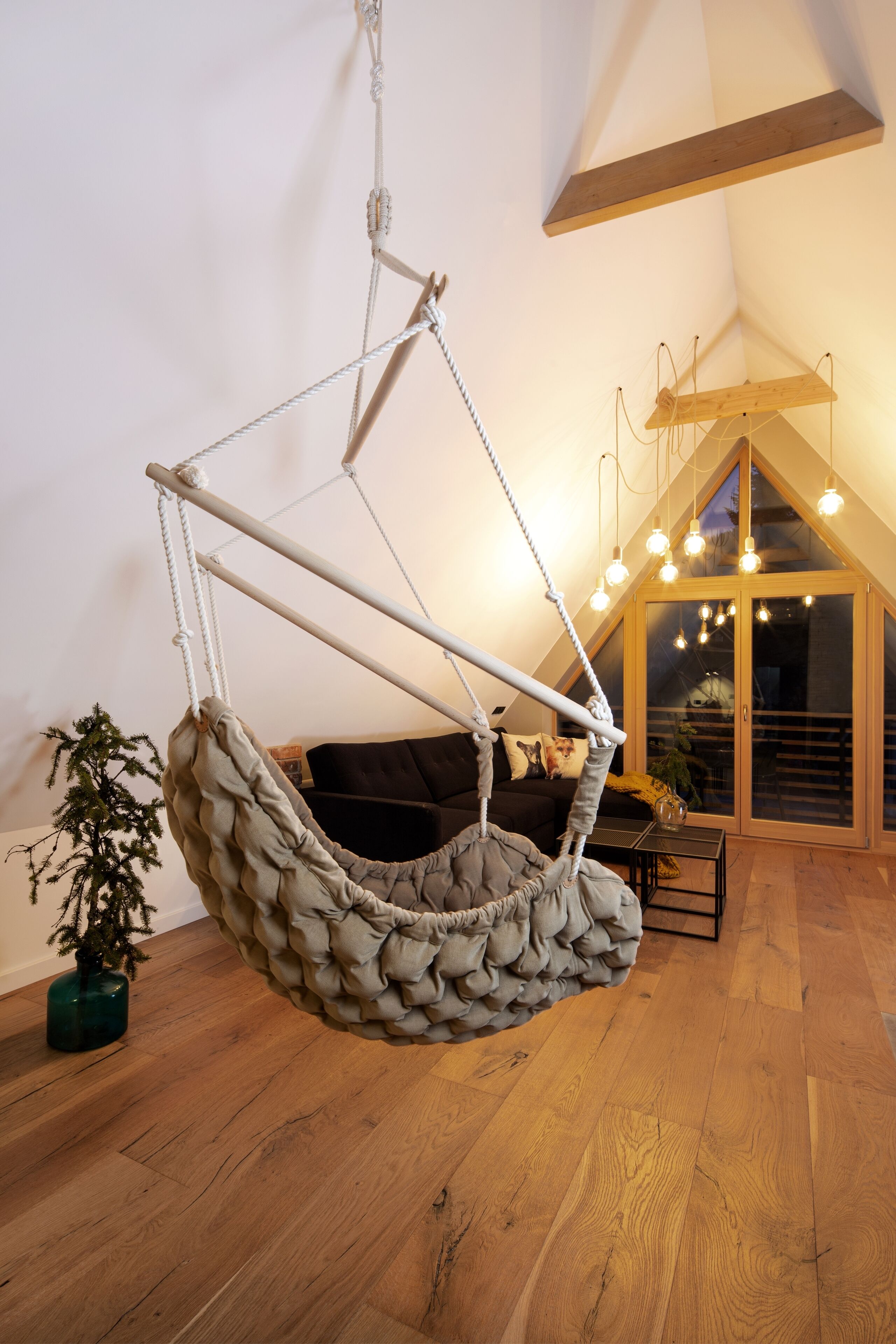 A cozy room with a hanging chair, wooden flooring, a potted plant, and a large window with lights.