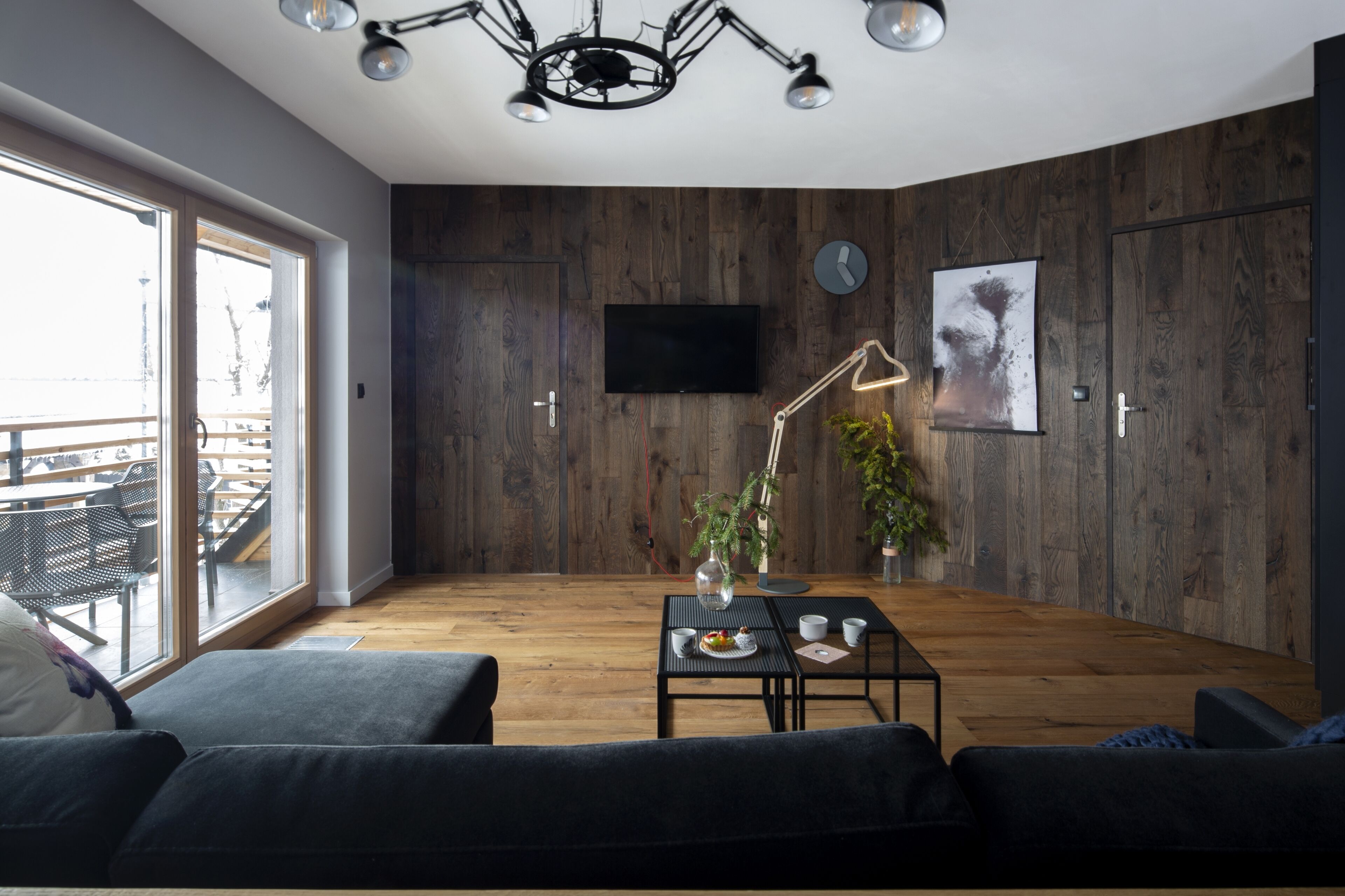 A modern living room with a wooden floor, a black sofa, a coffee table with a plant, a TV mounted on the wall, and a large window with a view of outdoor seating.