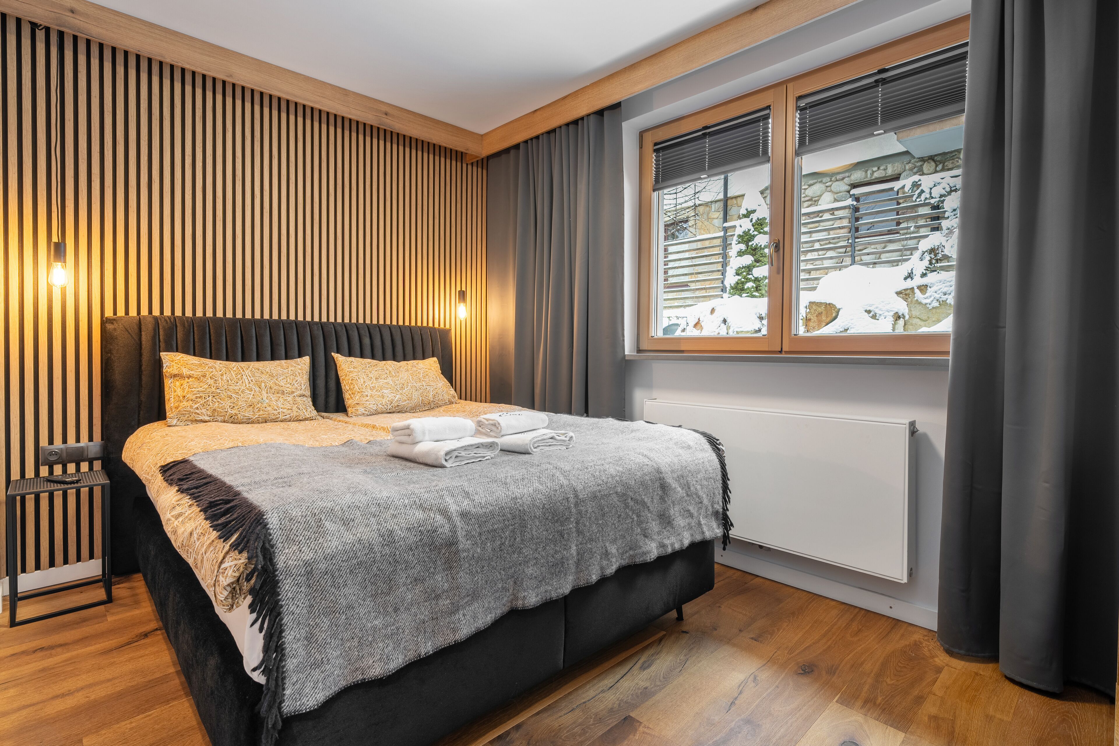 A bedroom with a bed, a bedside table, a window with a view of a snowy landscape, and a wall with vertical wooden slats.