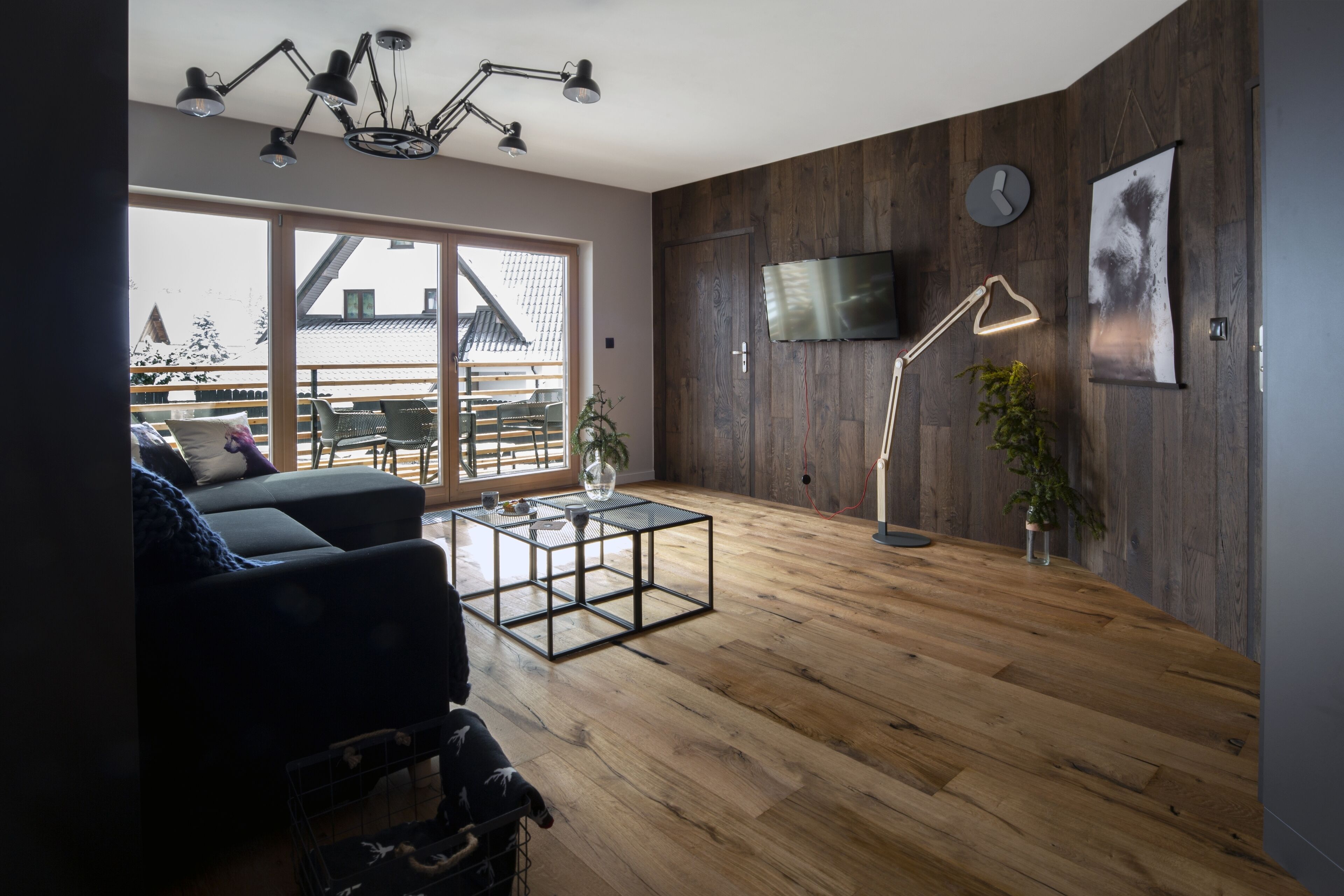A modern living room with a wooden floor, a black sofa, a glass coffee table, and a TV mounted on the wall.