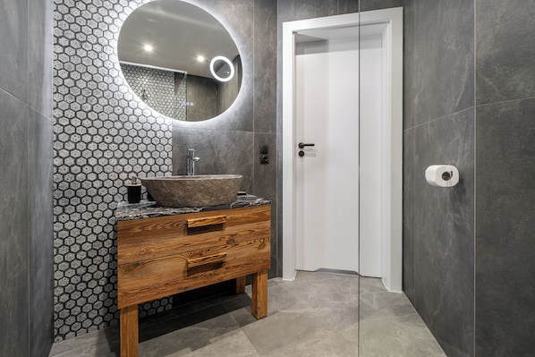 Un baño con tocador de madera, espejo redondo y salpicadero de azulejos hexagonales.