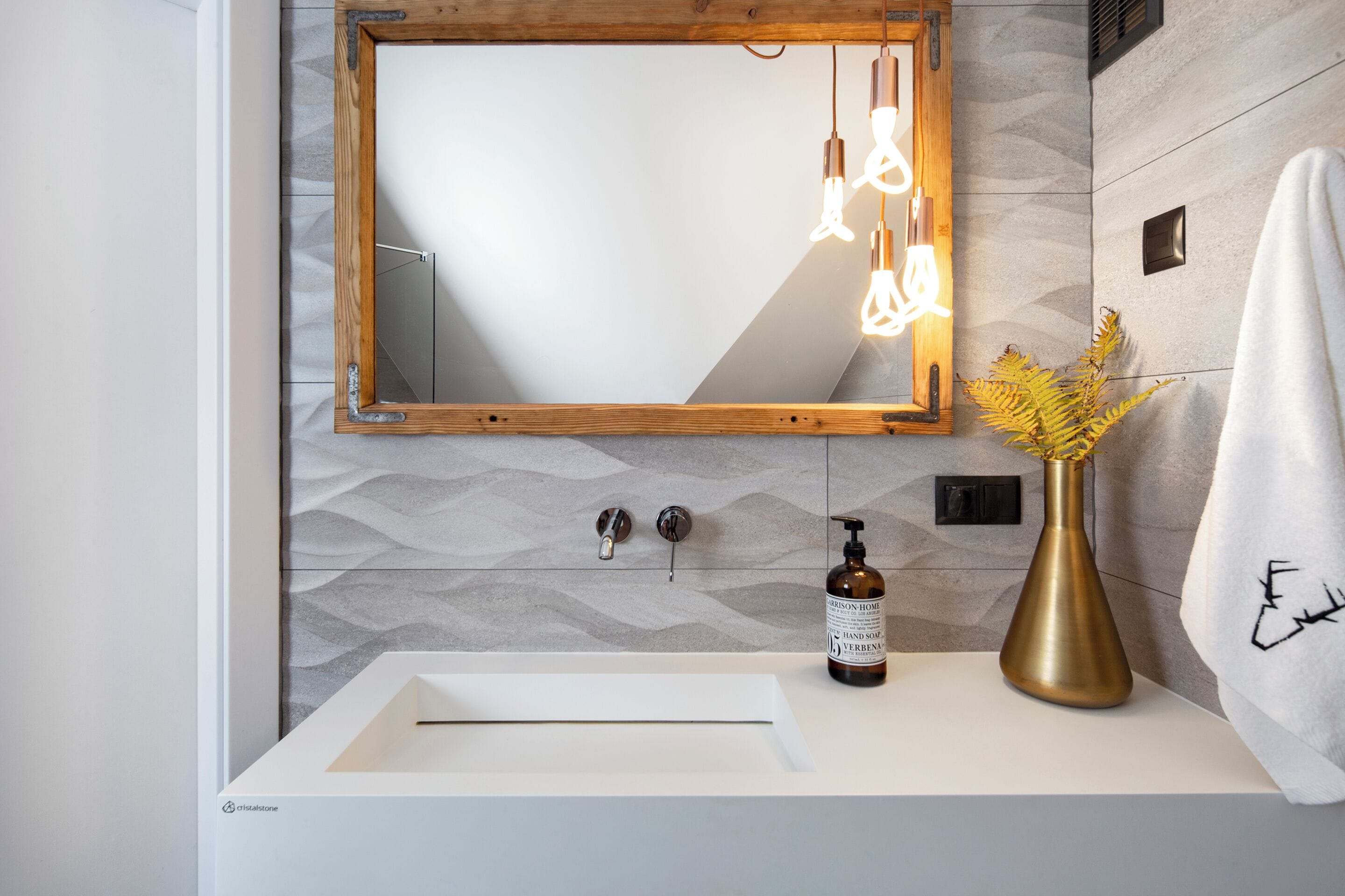 A bathroom with a white sink, a wooden-framed mirror, and a gold vase with yellow flowers.