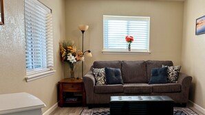 Un salon avec un canapé marron, un pouf noir, un tapis à motifs, une petite table d’appoint en bois sur laquelle est posé un vase de fleurs, et une fenêtre avec des stores.