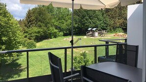 Un balcon avec une table et des chaises, un grand parasol et une vue sur un parc avec des arbres et une aire de jeux.