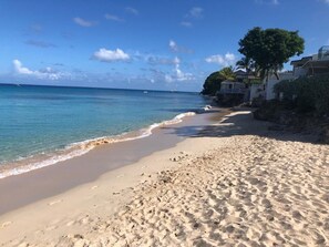 Une plage de sable fin bordée d’une eau d’un bleu limpide, quelques empreintes de pas et un bâtiment avec un arbre sur la droite.