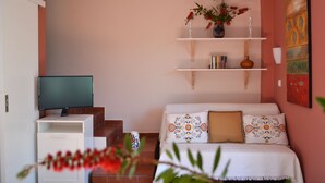 Ett vardagsrum med en tv på ett tv-bord, en soffa med mönstrade kuddar, en bokhylla med böcker och en vas med blommor samt en tavla på väggen.