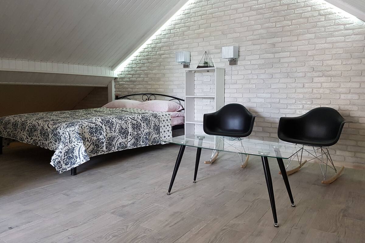 Un dormitorio con techo inclinado, una cama con ropa de cama estampada, una mesita de noche, dos sillas negras y una pared de ladrillo blanco.