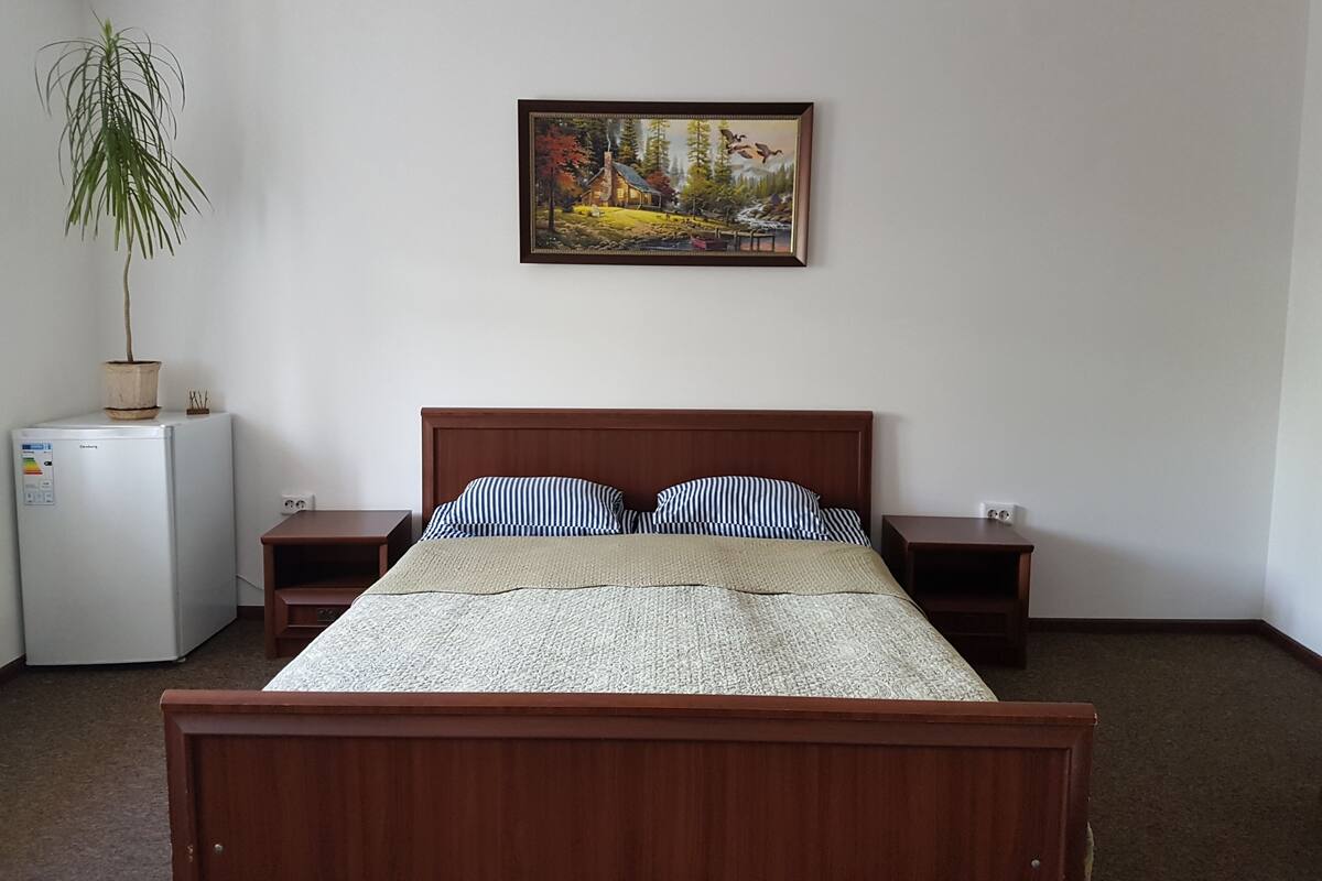 Un dormitorio con una cama de madera, dos mesitas de noche, un refrigerador blanco pequeño y una planta en maceta.