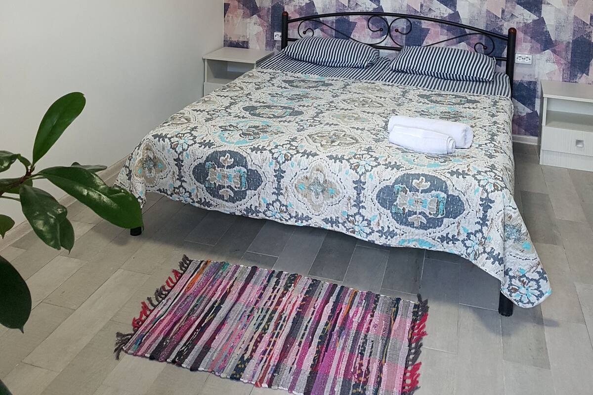 Un dormitorio con colcha estampada, una mesita de noche y una planta en maceta. En la pared, un diseño geométrico.