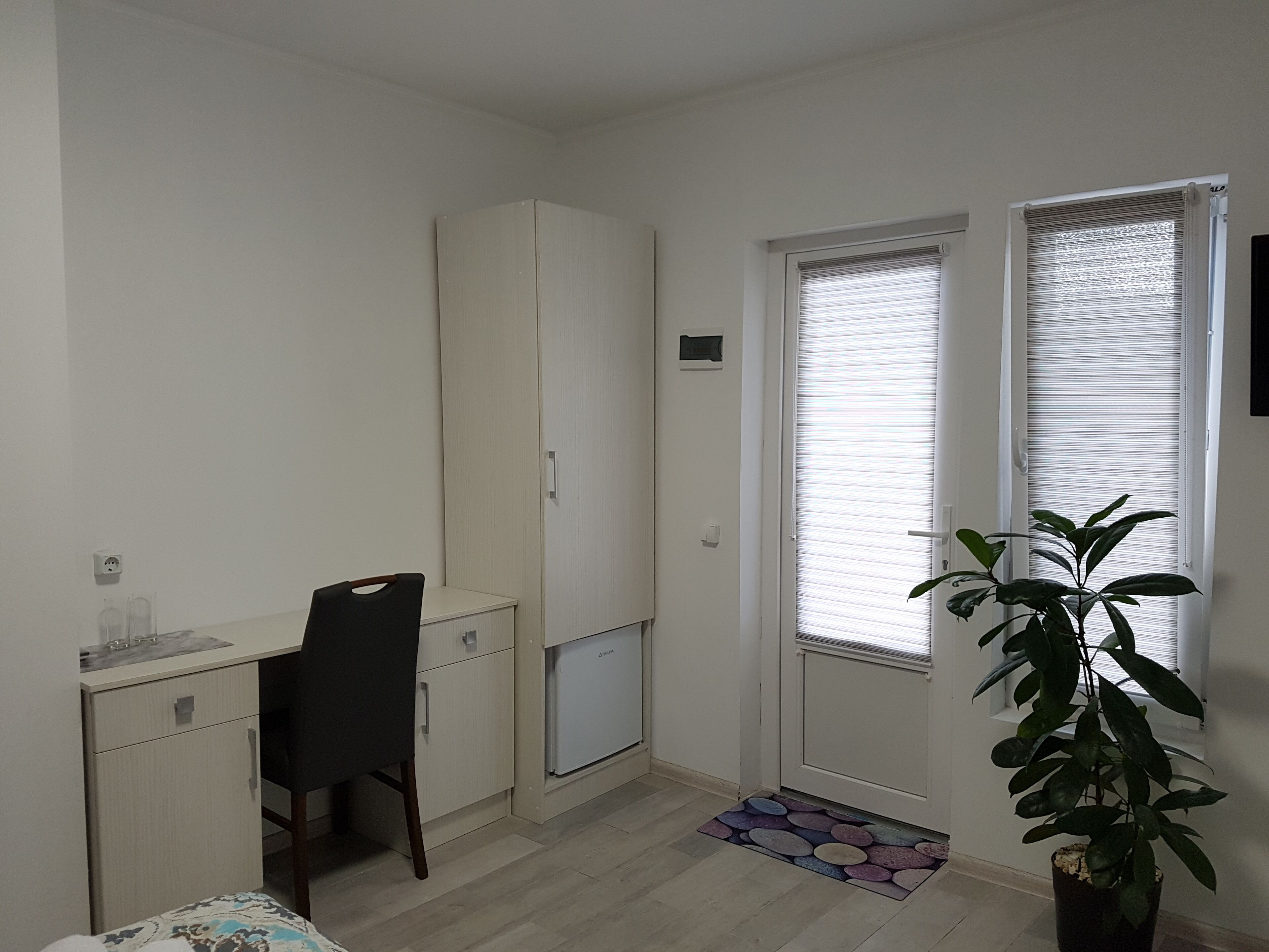 A small, sparsely furnished room with a desk, chair, wardrobe, and a potted plant.