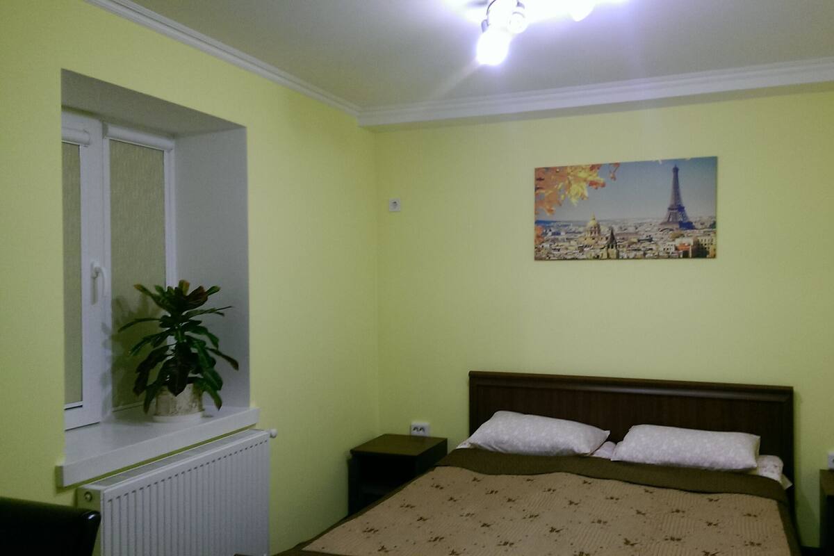 Un dormitorio con una cama, dos mesitas de noche, una ventana con una planta y un cuadro de un paisaje urbano.