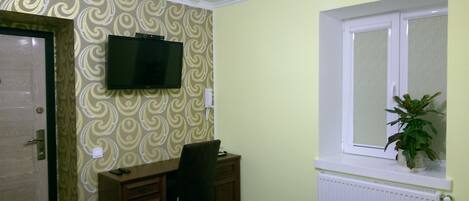 Una habitación con papel tapiz estampado, un televisor montado, un escritorio con una silla, una ventana con una planta y un radiador.