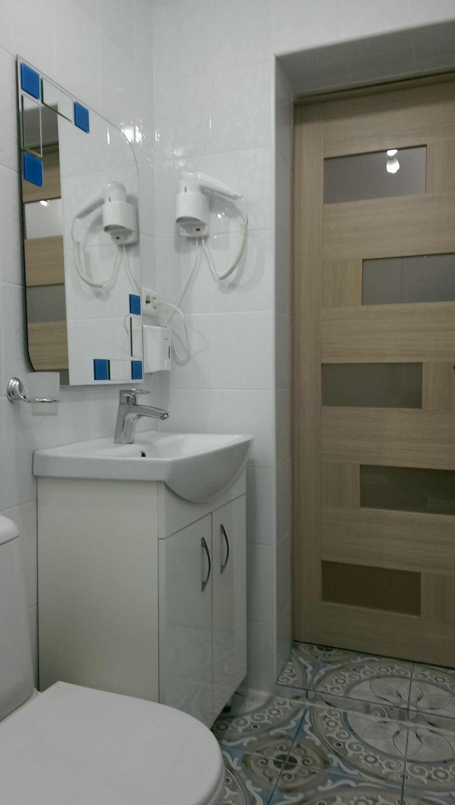 A bathroom with a white sink, mirror, and toilet. There is a wooden door with glass panels and patterned floor tiles.