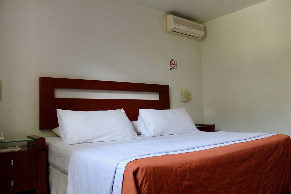 A neatly made bed with white linens and an orange blanket, a wooden headboard, a bedside table with a lamp, and a wall-mounted air conditioner.