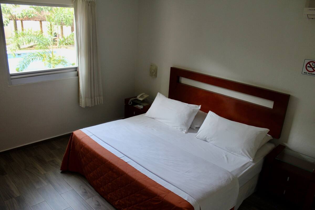 A neatly made bed with white linens and a brown bedspread in a hotel room with a wooden floor, a nightstand with a phone, and a window with curtains.