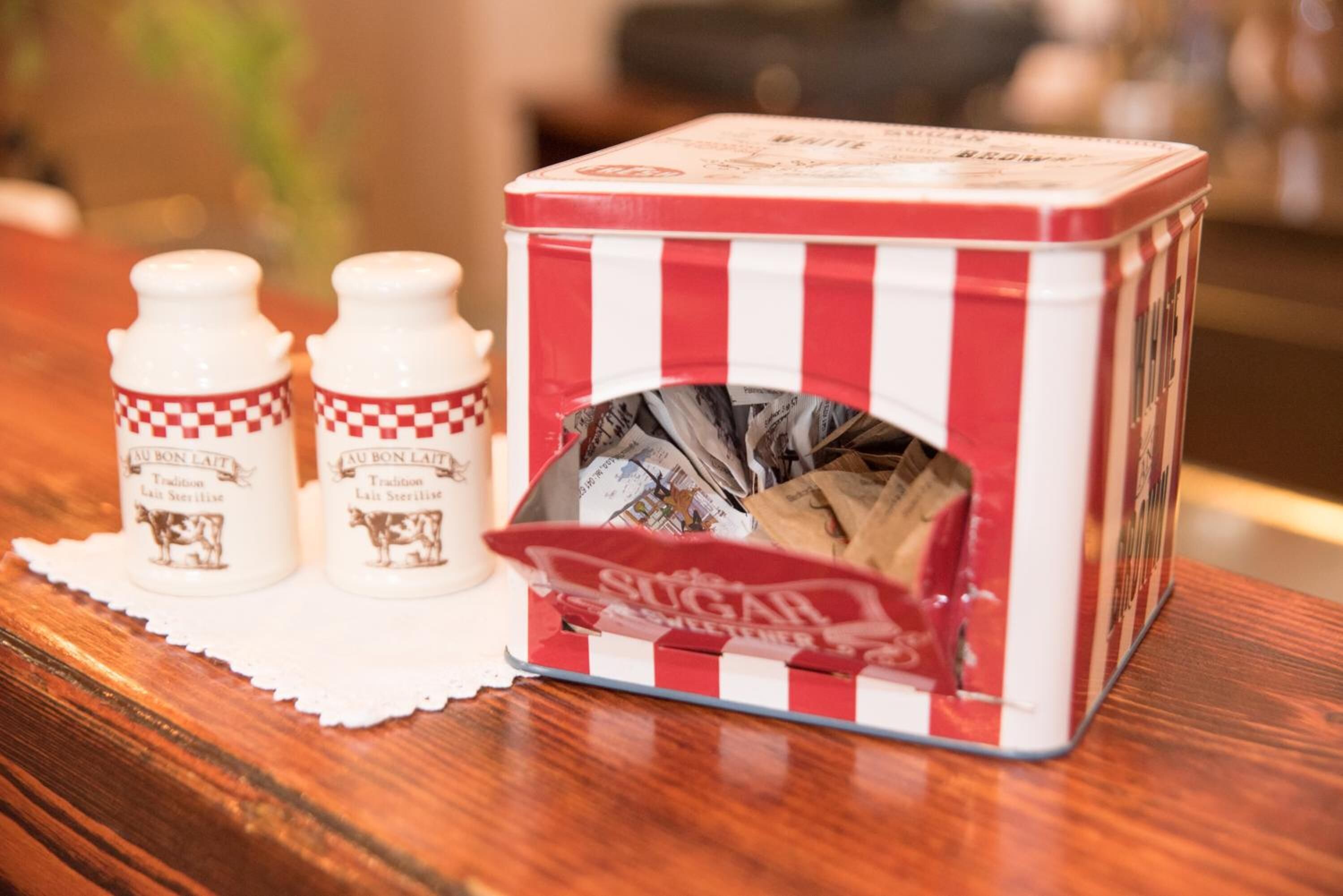A tin box with a red and white checkered pattern, containing sugar packets and two bottles labeled "LA BON L'ATT" with a cow image.