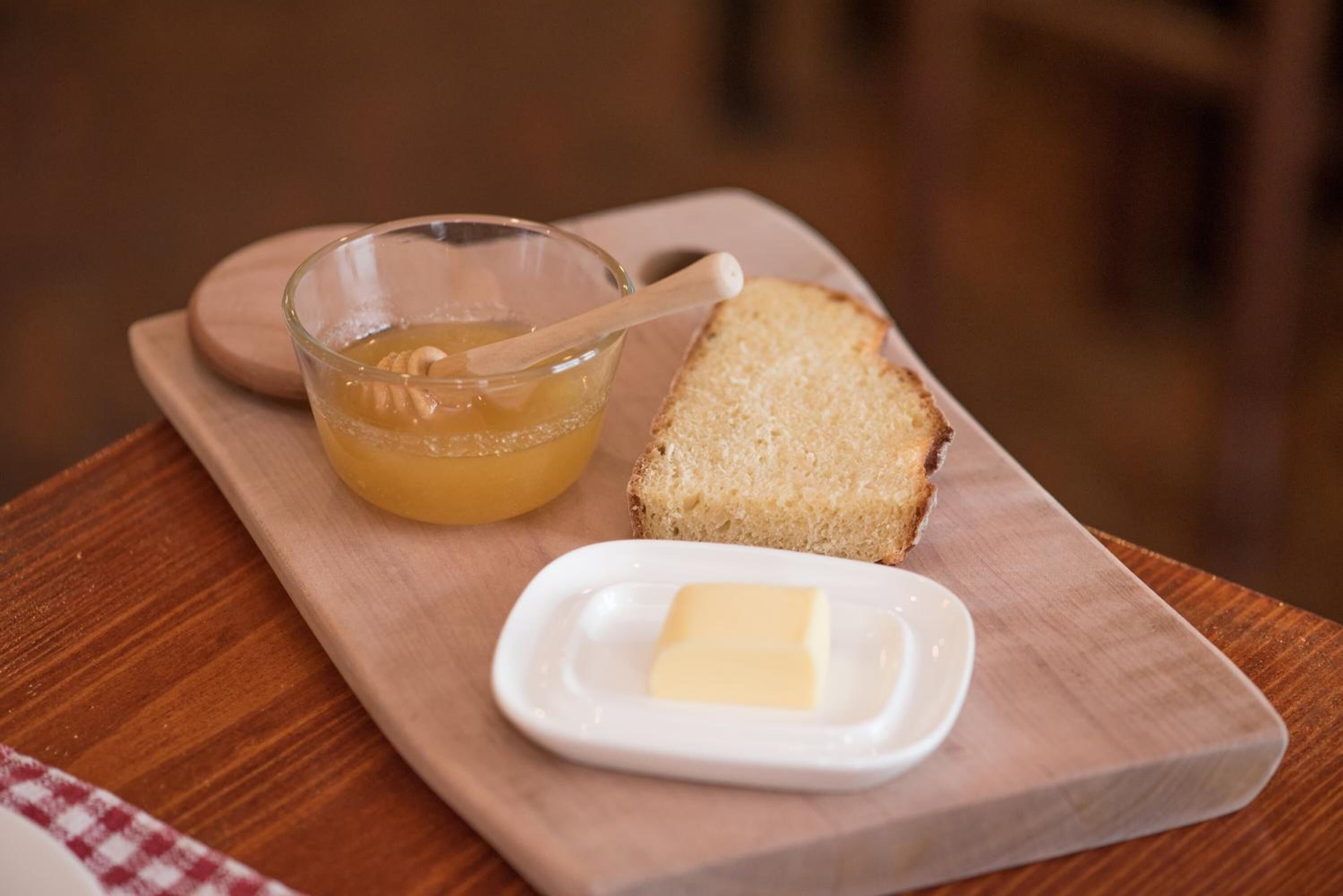 A wooden serving board with a glass bowl of honey, a piece of butter, and a slice of bread.